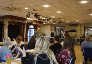 People sitting down observing a talk from a Dr and researcher from the University of Sheffield in a theatre-like setting.