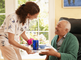 Carer interacting with a service user and handing him a tray with food and drink while he sits on a chair wearing a dark green dressing gown.