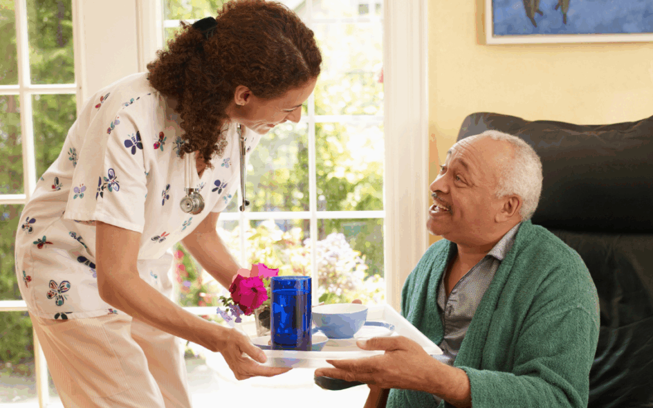 Carer interacting with a service user and handing him a tray with food and drink while he sits on a chair wearing a dark green dressing gown.
