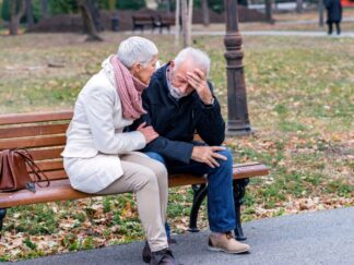 Depression in older adults representation: Two older adults sitting on a park bench outside. One man and one woman, presumably a married couple. Man has his head in his hands, looking distressed and woman is comforting him.